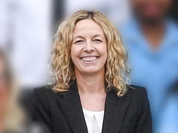 Portrait of a smiling woman with curly blonde hair wearing a black blazer and white top, standing in front of a blurred team background.