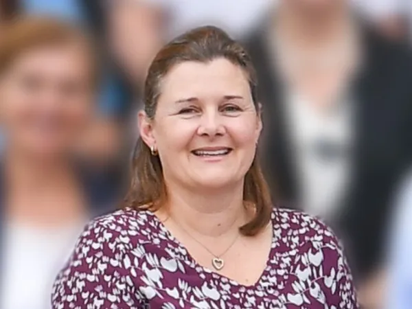 Portrait of a smiling woman in a purple patterned blouse, standing in front of a blurred group background.