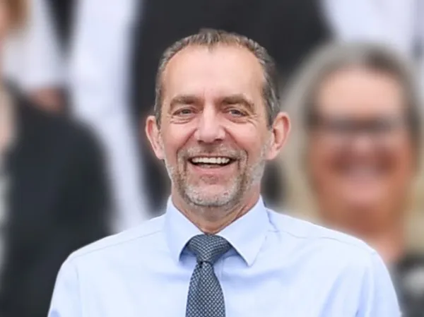 Smiling man in a light blue shirt and patterned tie, standing in front of a blurred team background.