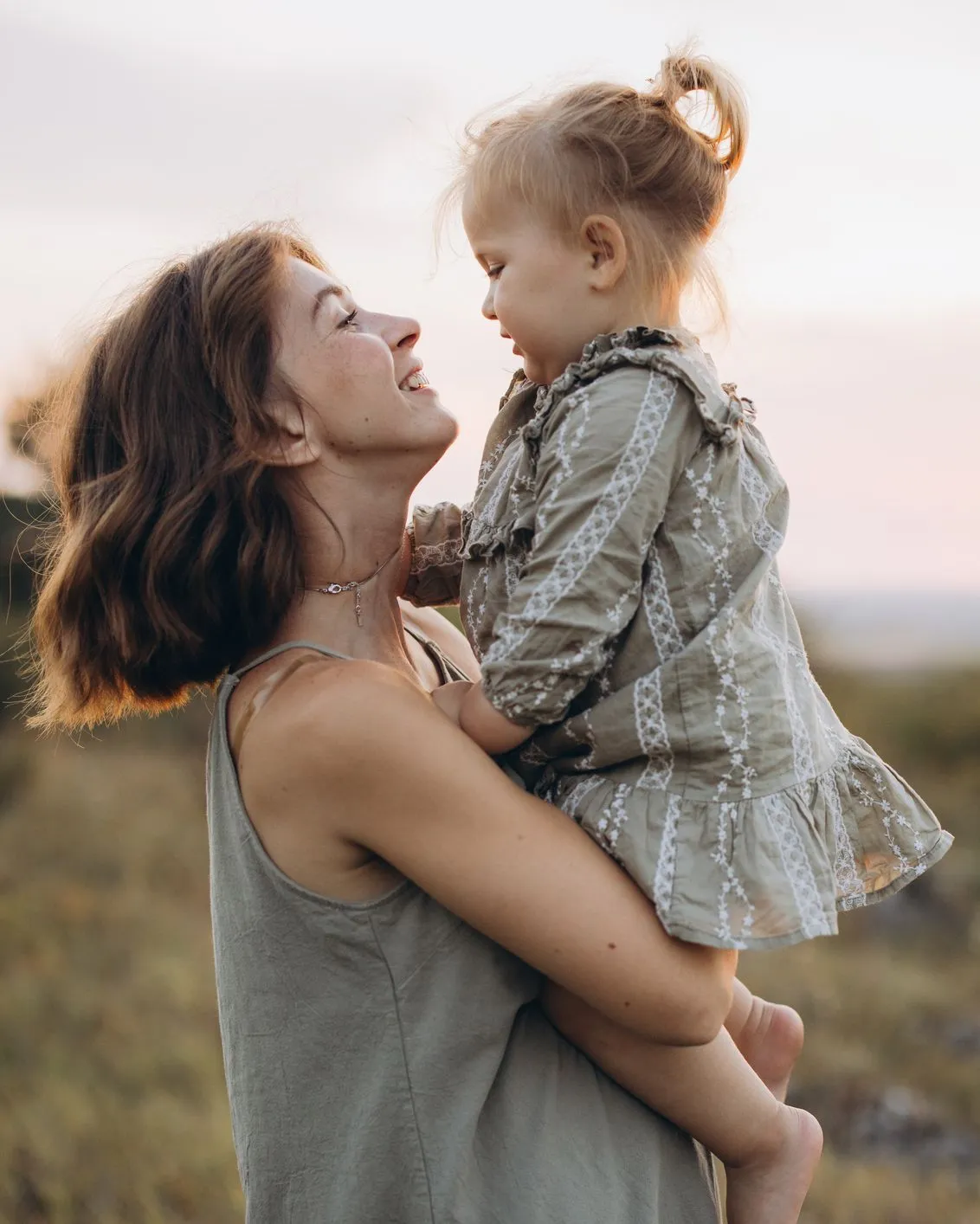 Mother holding her young daughter outdoors at sunset, smiling at each other