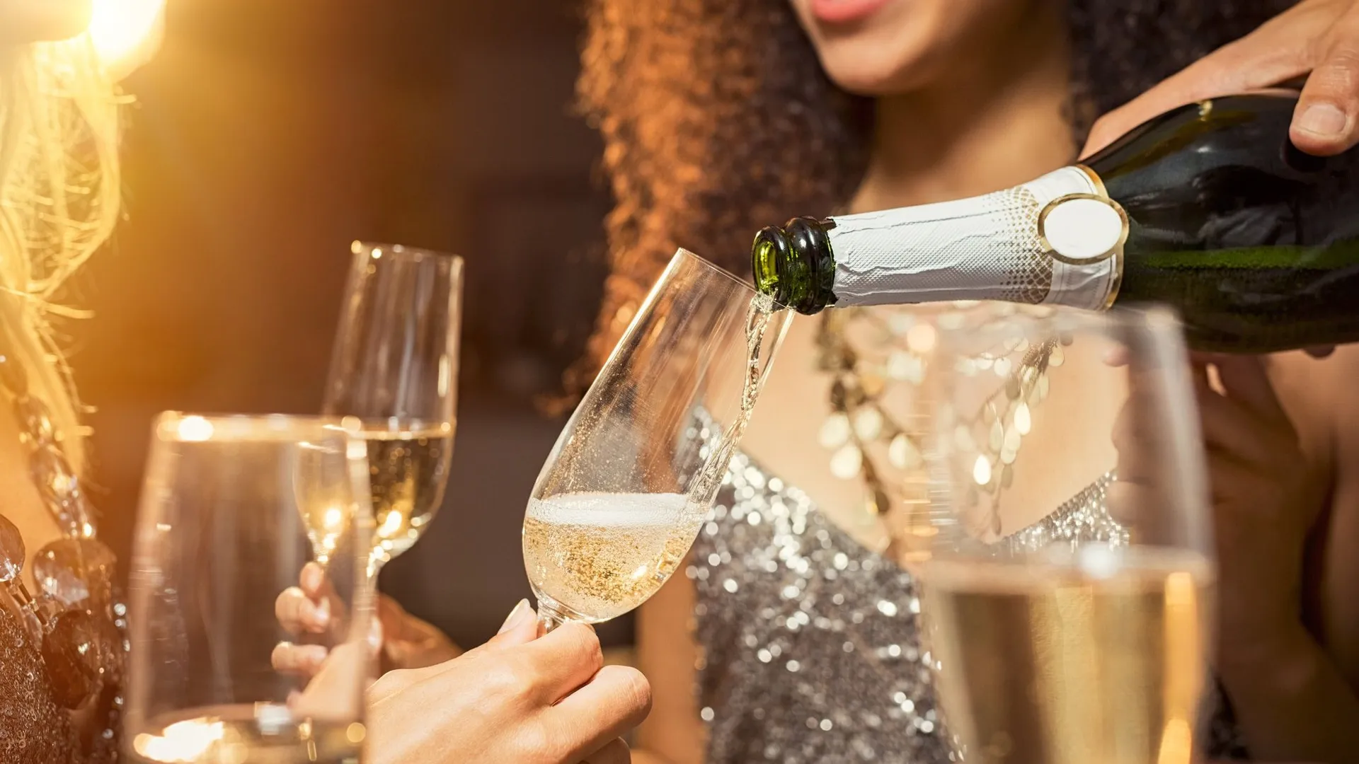 Sparkling wine being poured into a flute at a New Year’s Eve party, with smiling guests in festive attire holding glasses.