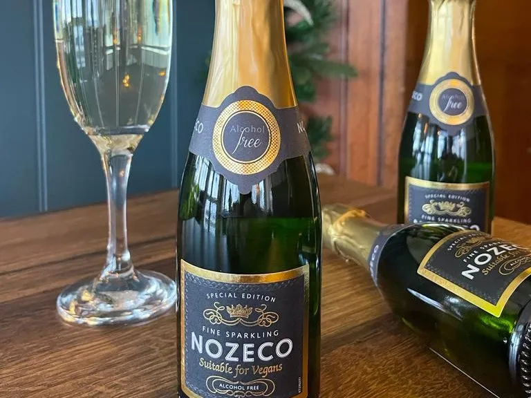 Bottle of Nozeco alcohol-free sparkling drink beside a filled flute glass, with two more bottles on a wooden table and festive decorations in the background