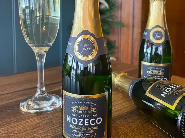 Bottle of Nozeco alcohol-free sparkling drink beside a filled flute glass, with two more bottles on a wooden table and festive decorations in the background