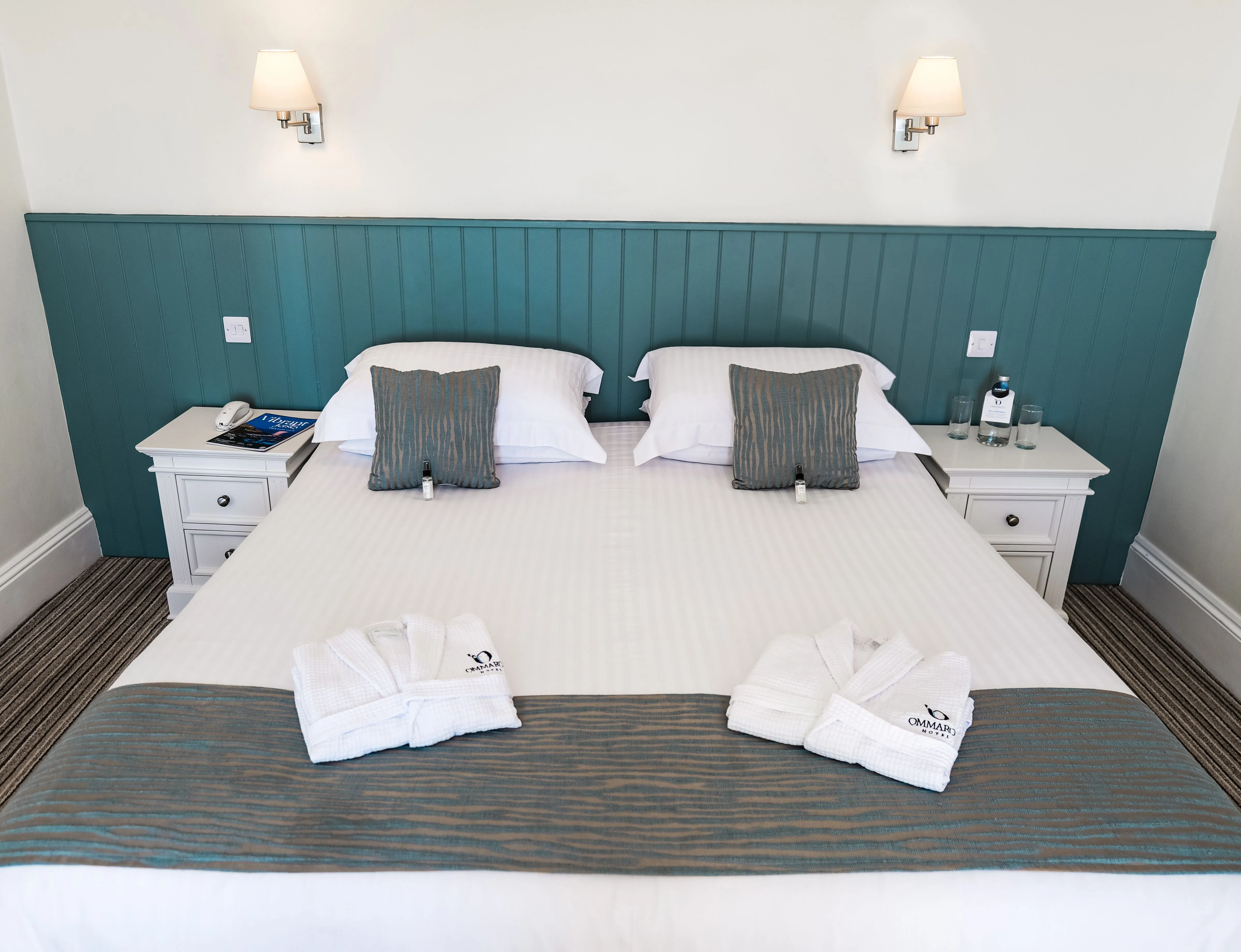 Neatly made double bed in a bright hotel room with teal panelled headboard, white bedside tables and folded bathrobes.