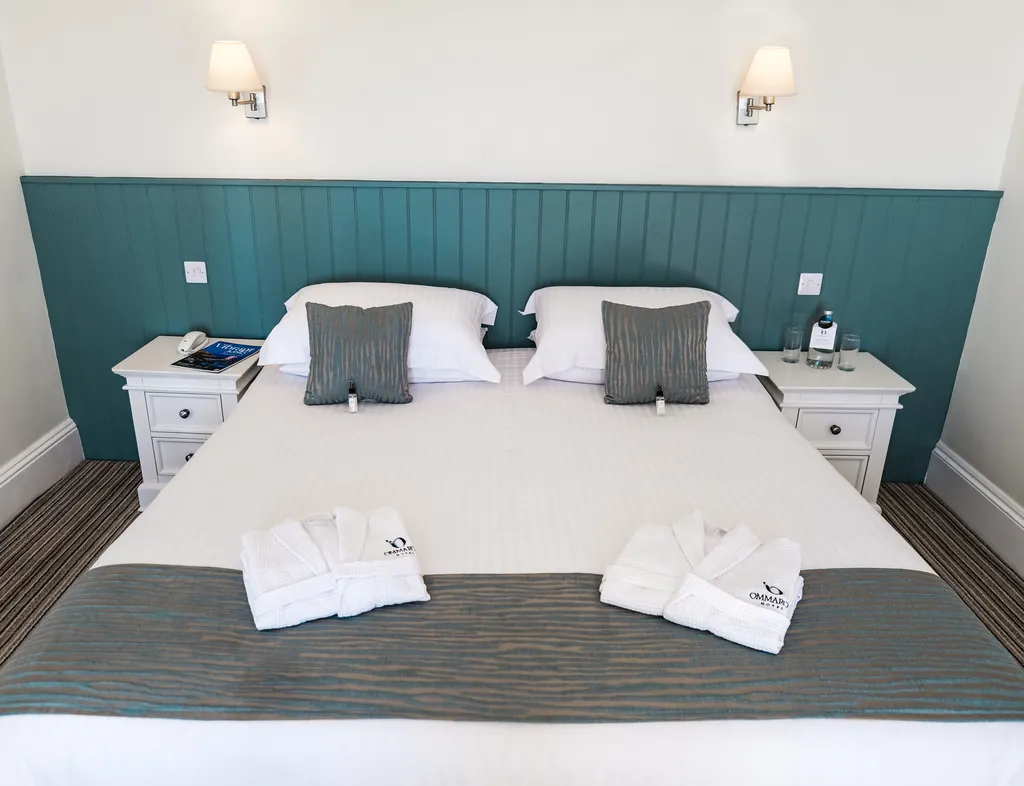 Neatly made double bed in a bright hotel room with teal panelled headboard, white bedside tables and folded bathrobes.