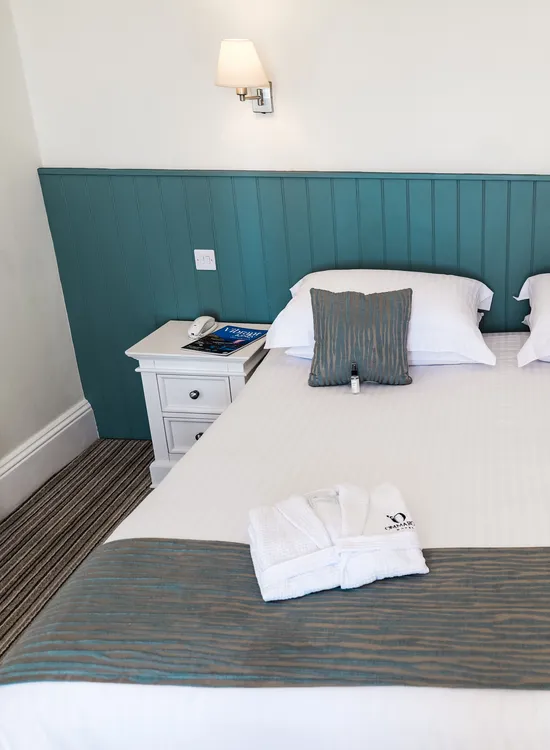 Neatly made double bed in a bright hotel room with teal panelled headboard, white bedside tables and folded bathrobes.