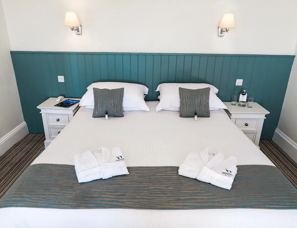 Neatly made double bed in a bright hotel room with teal panelled headboard, white bedside tables and folded bathrobes.