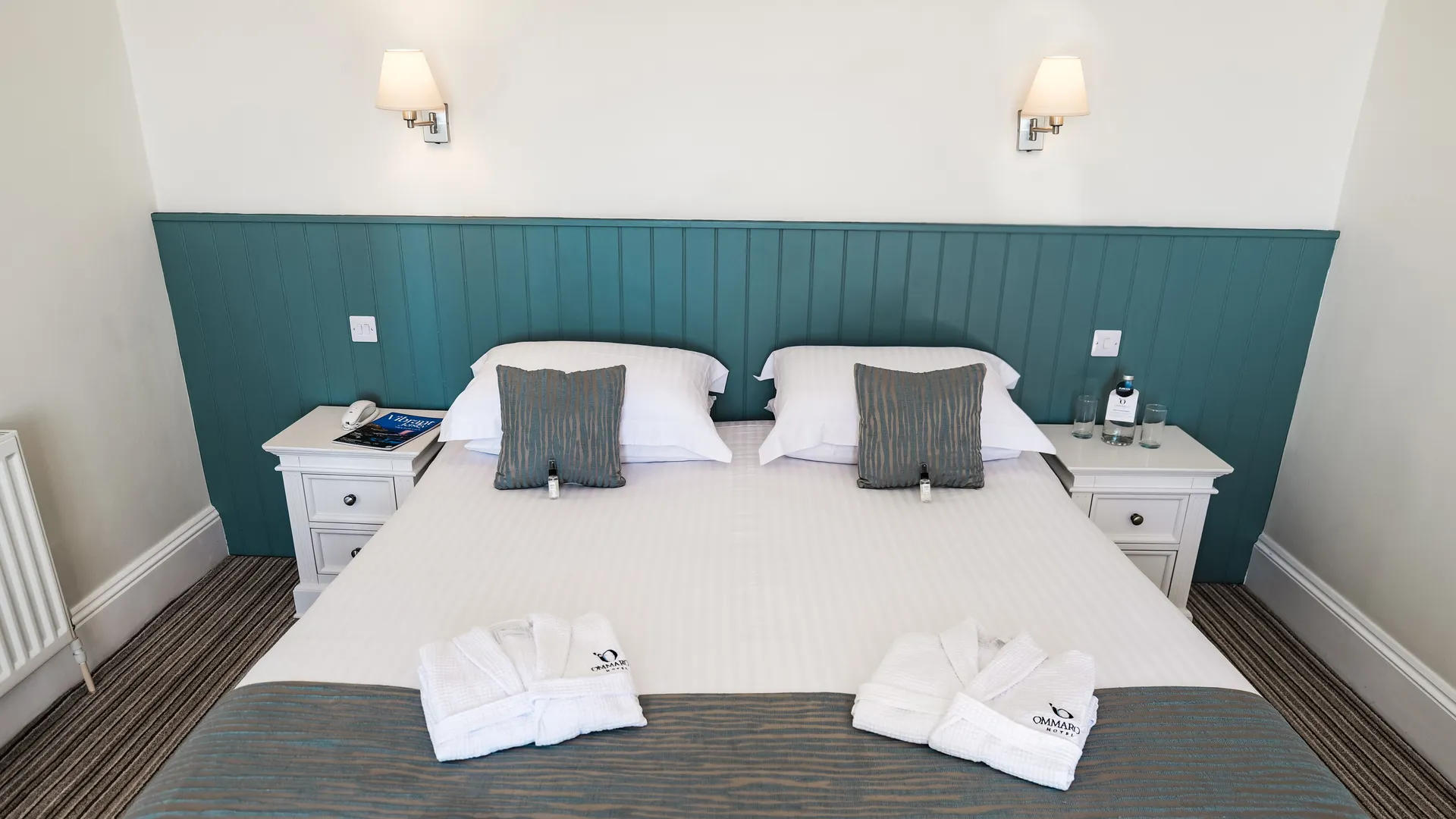 Neatly made double bed in a bright hotel room with teal panelled headboard, white bedside tables and folded bathrobes.