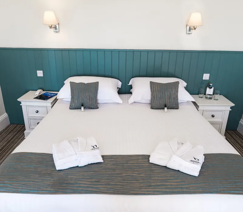 Neatly made double bed in a bright hotel room with teal panelled headboard, white bedside tables and folded bathrobes.