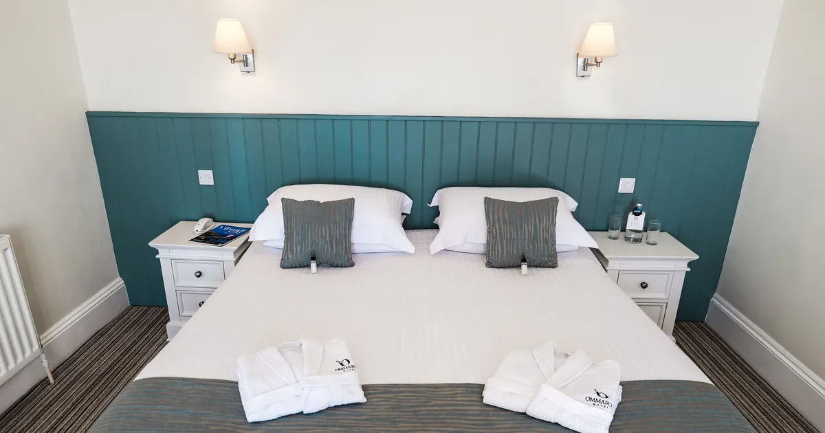 Neatly made double bed in a bright hotel room with teal panelled headboard, white bedside tables and folded bathrobes.