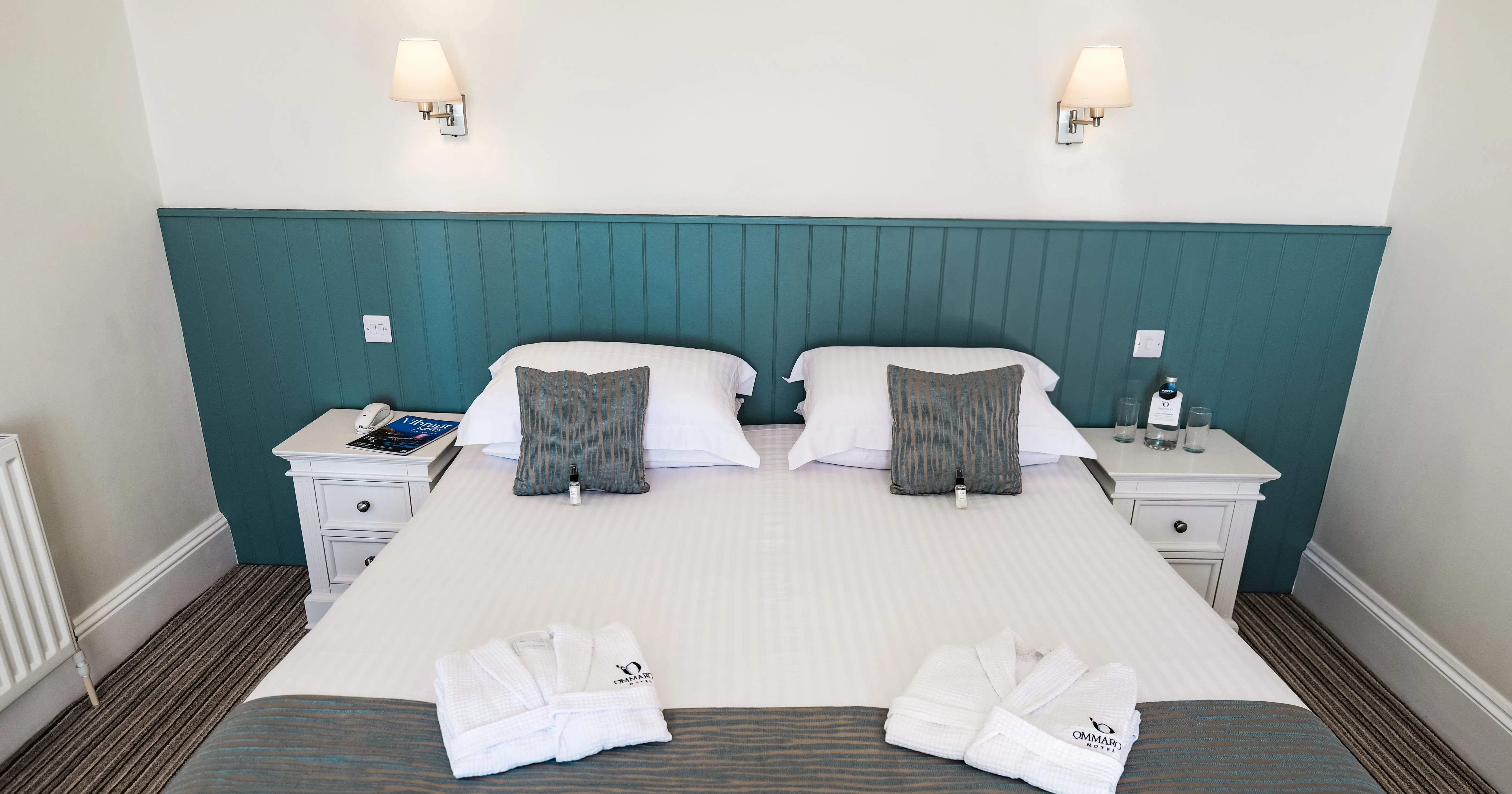 Neatly made double bed in a bright hotel room with teal panelled headboard, white bedside tables and folded bathrobes.