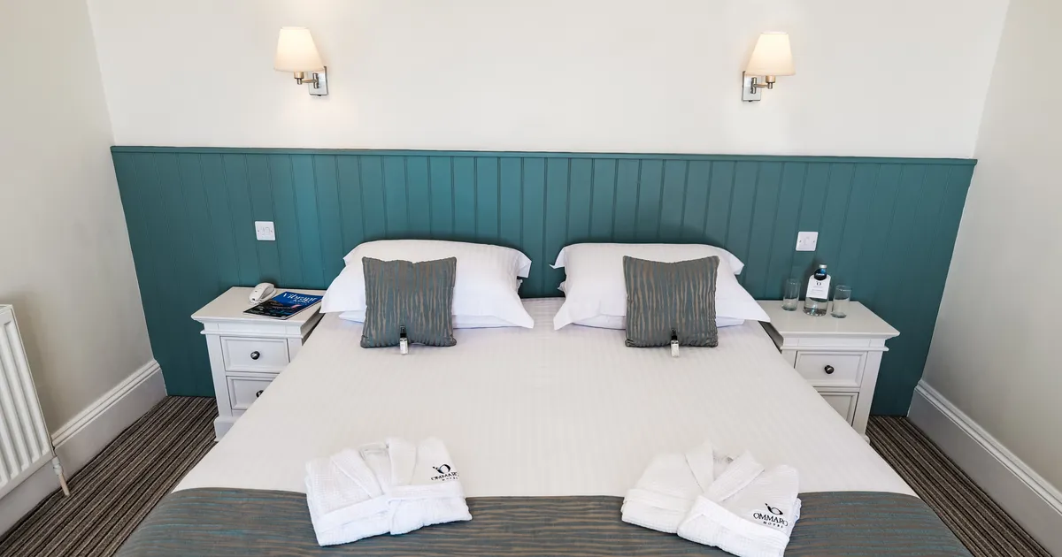 Neatly made double bed in a bright hotel room with teal panelled headboard, white bedside tables and folded bathrobes.