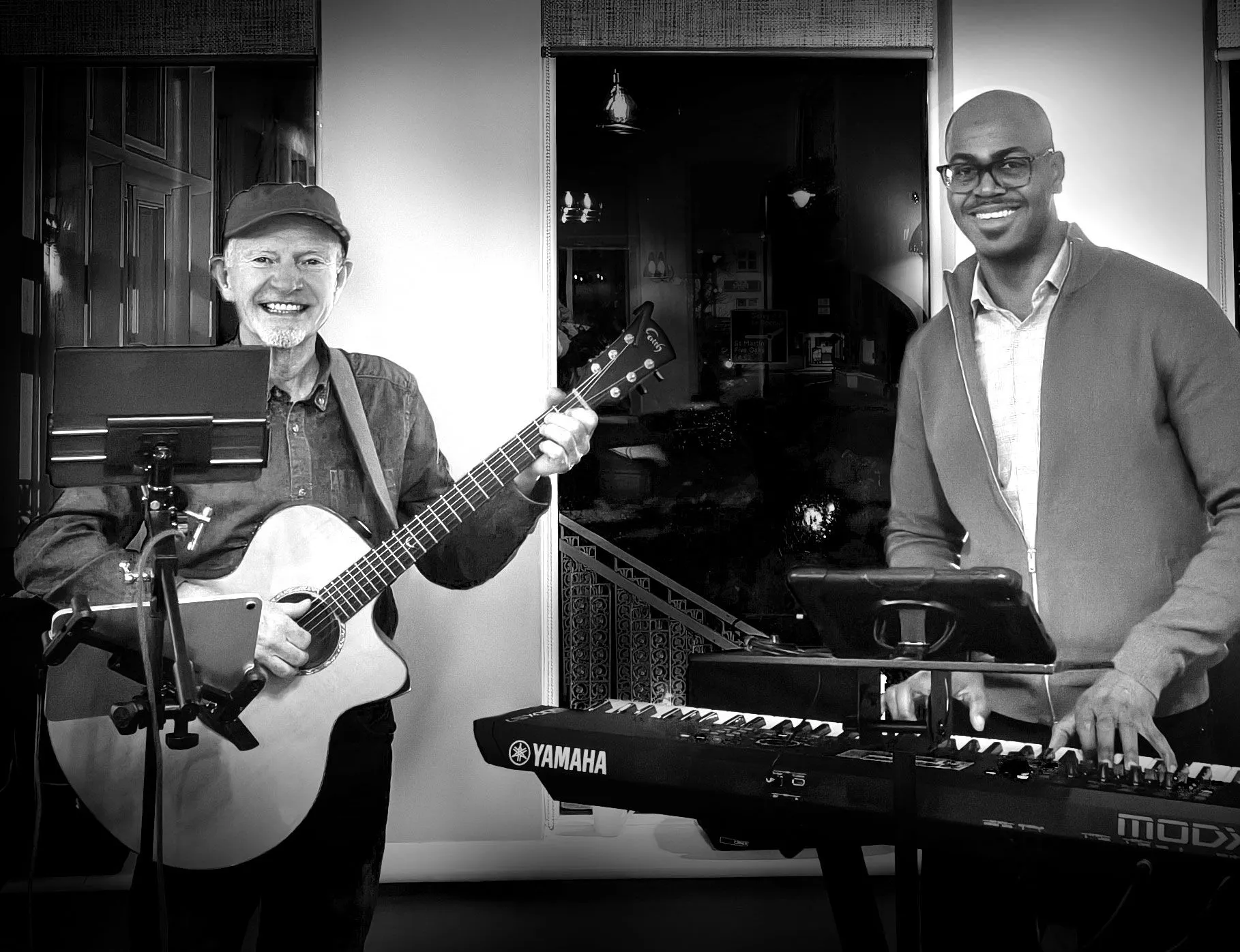 Black-and-white promotional image of two smiling musicians performing indoors, with one playing an acoustic guitar and the other standing at a keyboard.
