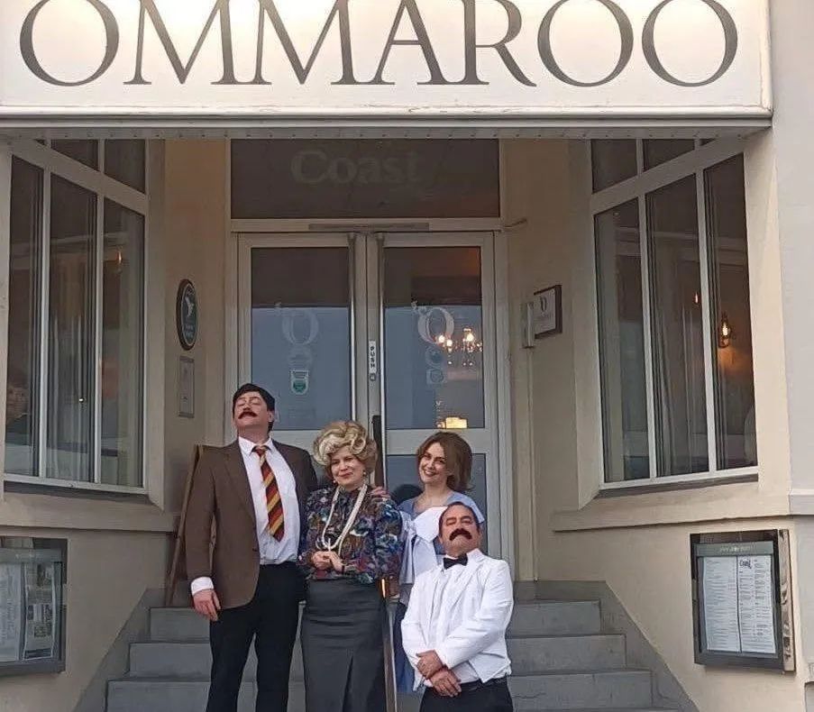 Four people in Fawlty Towers-inspired costumes posing on the steps beneath an OMMAROO hotel sign.