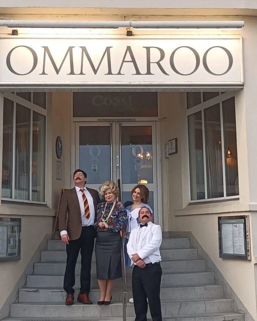 Four people in Fawlty Towers-inspired costumes posing on the steps beneath an OMMAROO hotel sign.