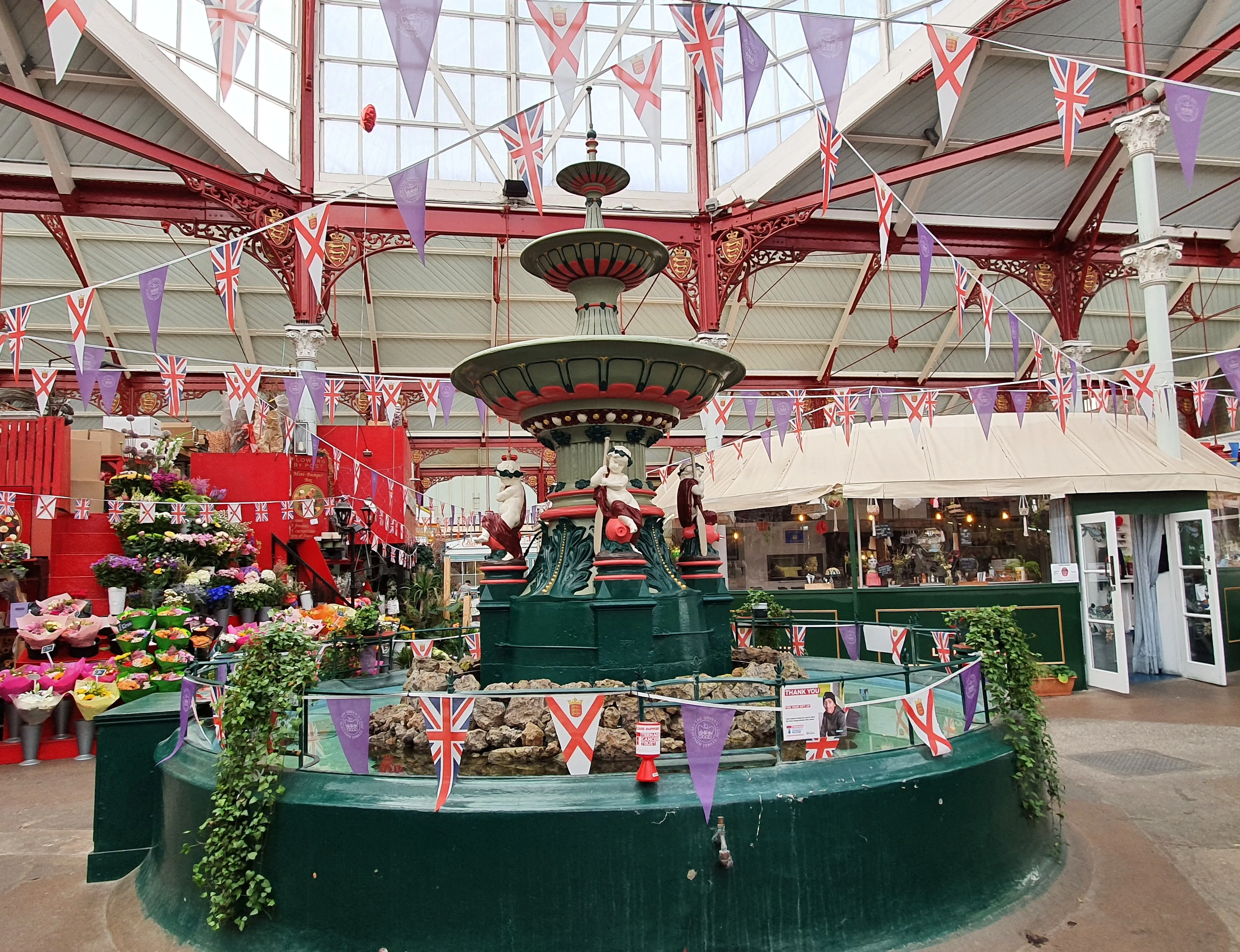 Ornate indoor market fountain decorated with Union Jack and red cross bunting, beside colourful flower stalls.