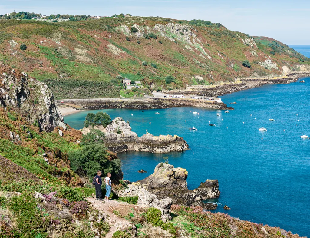 View over Bouley Bay with rocky cliffs, hillside vegetation, moored boats and two walkers on a coastal path.