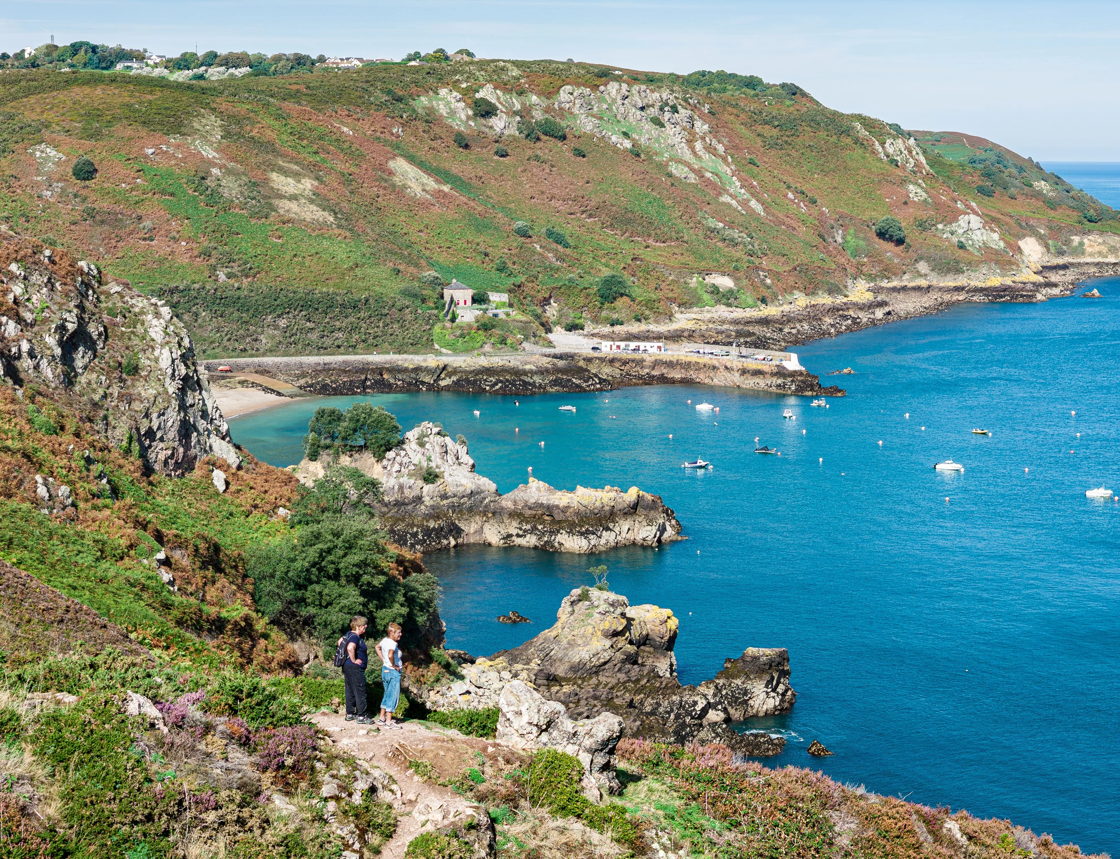 View over Bouley Bay with rocky cliffs, hillside vegetation, moored boats and two walkers on a coastal path.