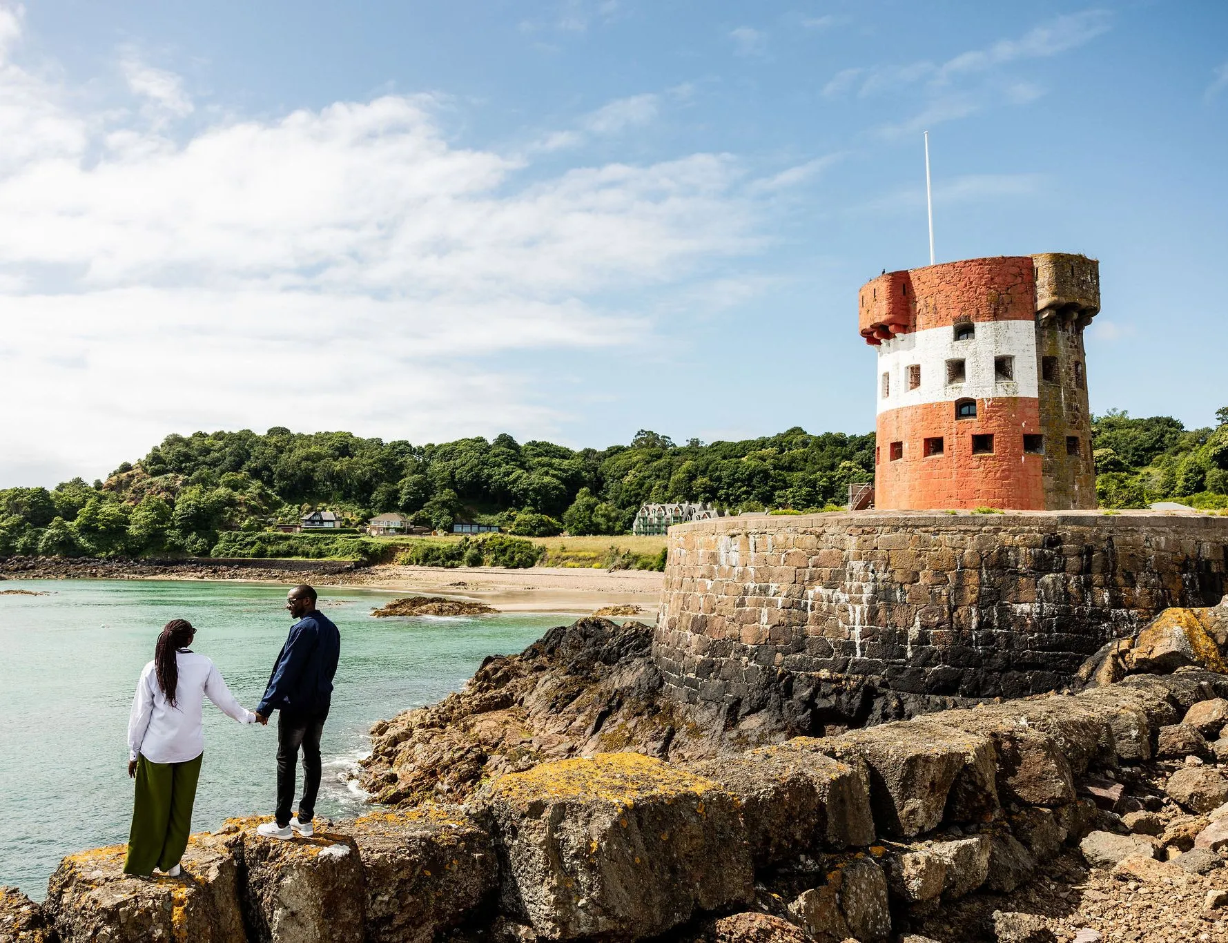 Couple standing on rocky shoreline beside Archirondel Tower overlooking turquoise water in Jersey.