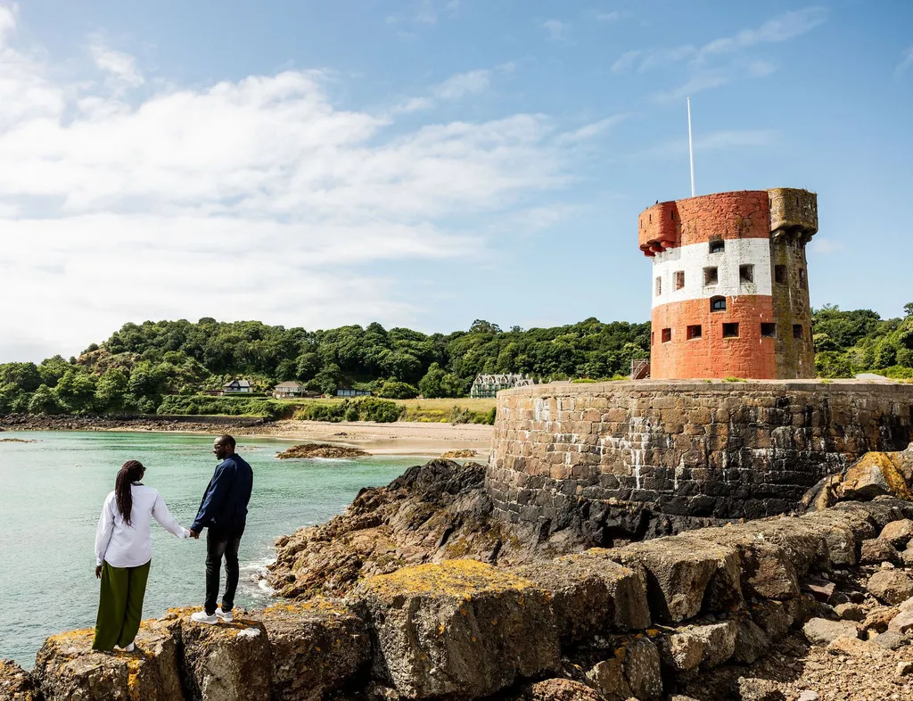 Couple standing on rocky shoreline beside Archirondel Tower overlooking turquoise water in Jersey.
