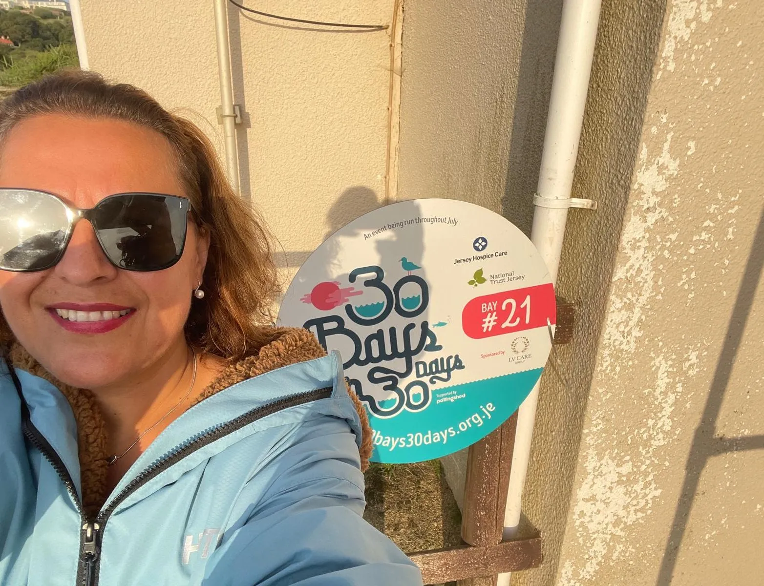 Smiling woman in sunglasses beside a 30 Bays in 30 Days Bay 21 charity event sign