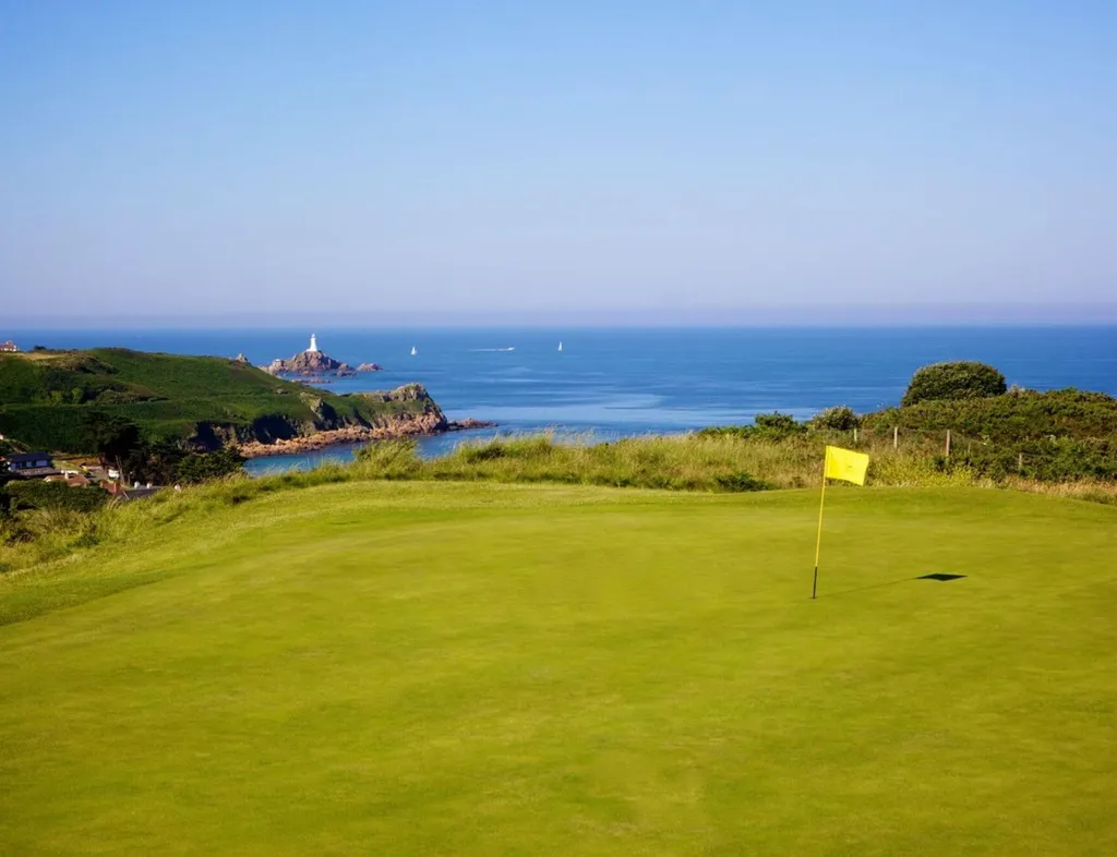 Golf putting green with a yellow flag overlooking a rocky coastline and lighthouse