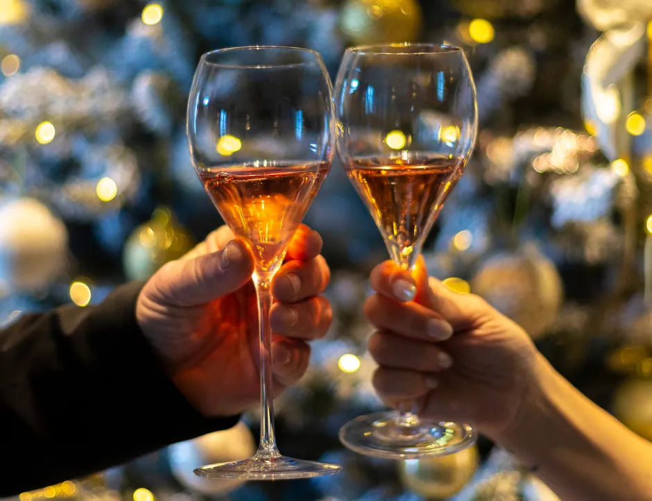 Two hands raising glasses of sparkling rosé in front of a softly lit Christmas tree.