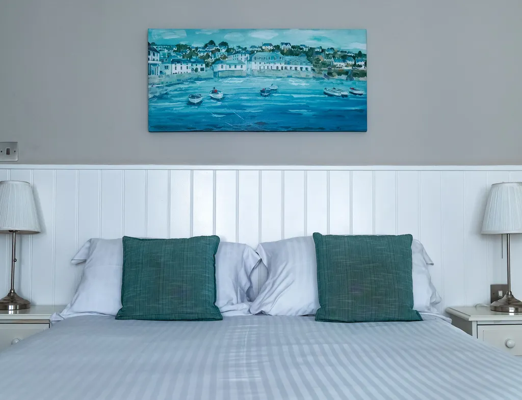 Hotel bedroom with a neatly made double bed, teal cushions, bedside lamps and coastal artwork.