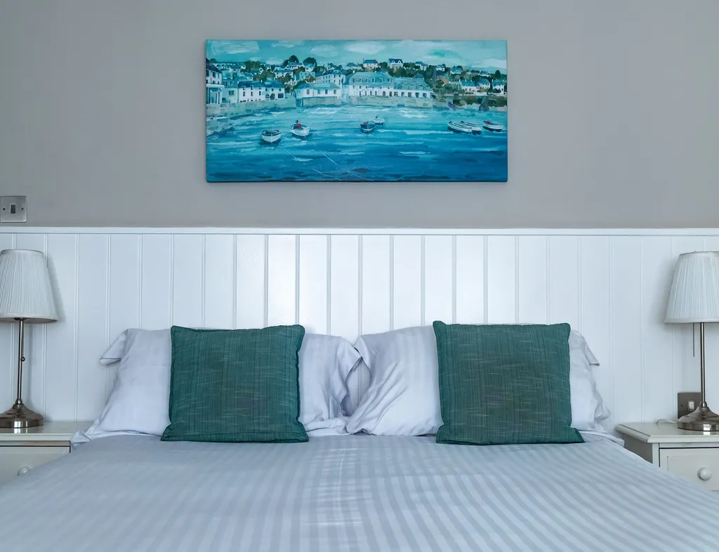 Hotel bedroom with a neatly made double bed, teal cushions, bedside lamps and coastal artwork.