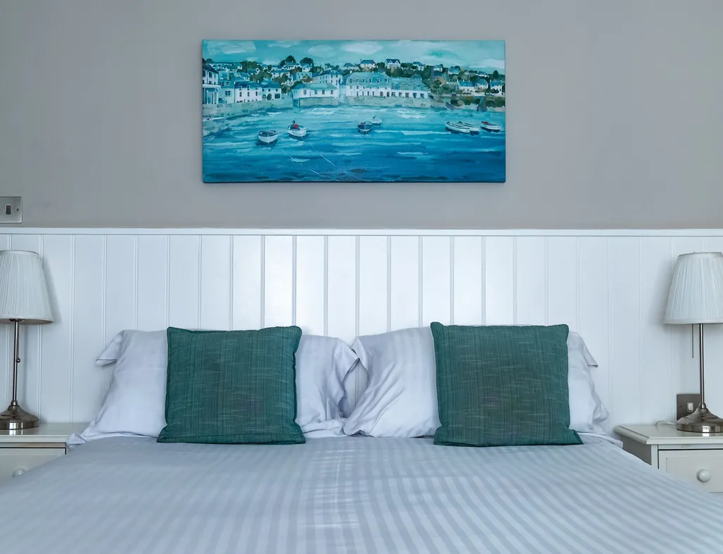 Hotel bedroom with a neatly made double bed, teal cushions, bedside lamps and coastal artwork.
