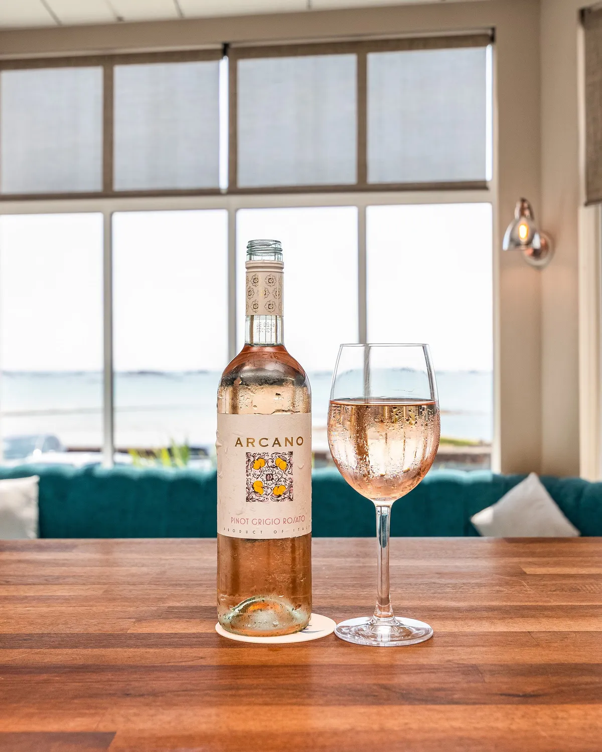 Bottle of rosé wine and a filled wine glass on a wooden table in a bright coastal bar