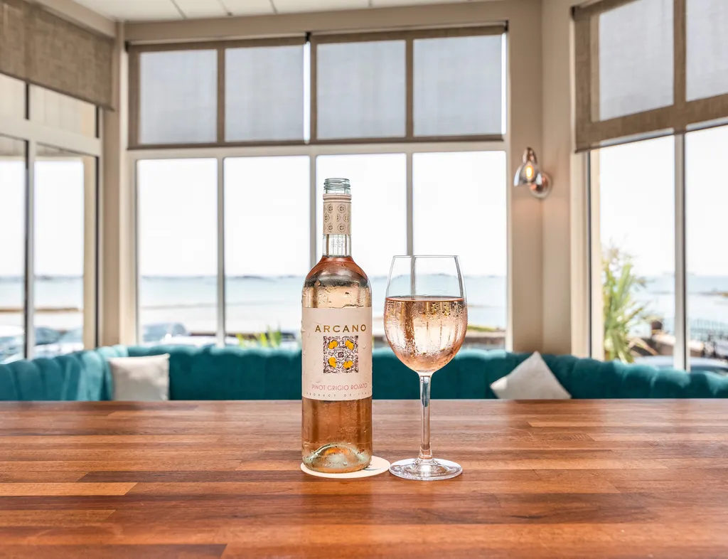 Bottle of rosé wine and a filled wine glass on a wooden table in a bright coastal bar