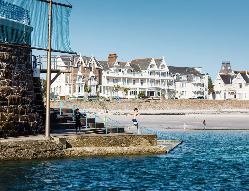 Boy jumping from a seaside harbour wall into clear blue water with beachfront buildings behind