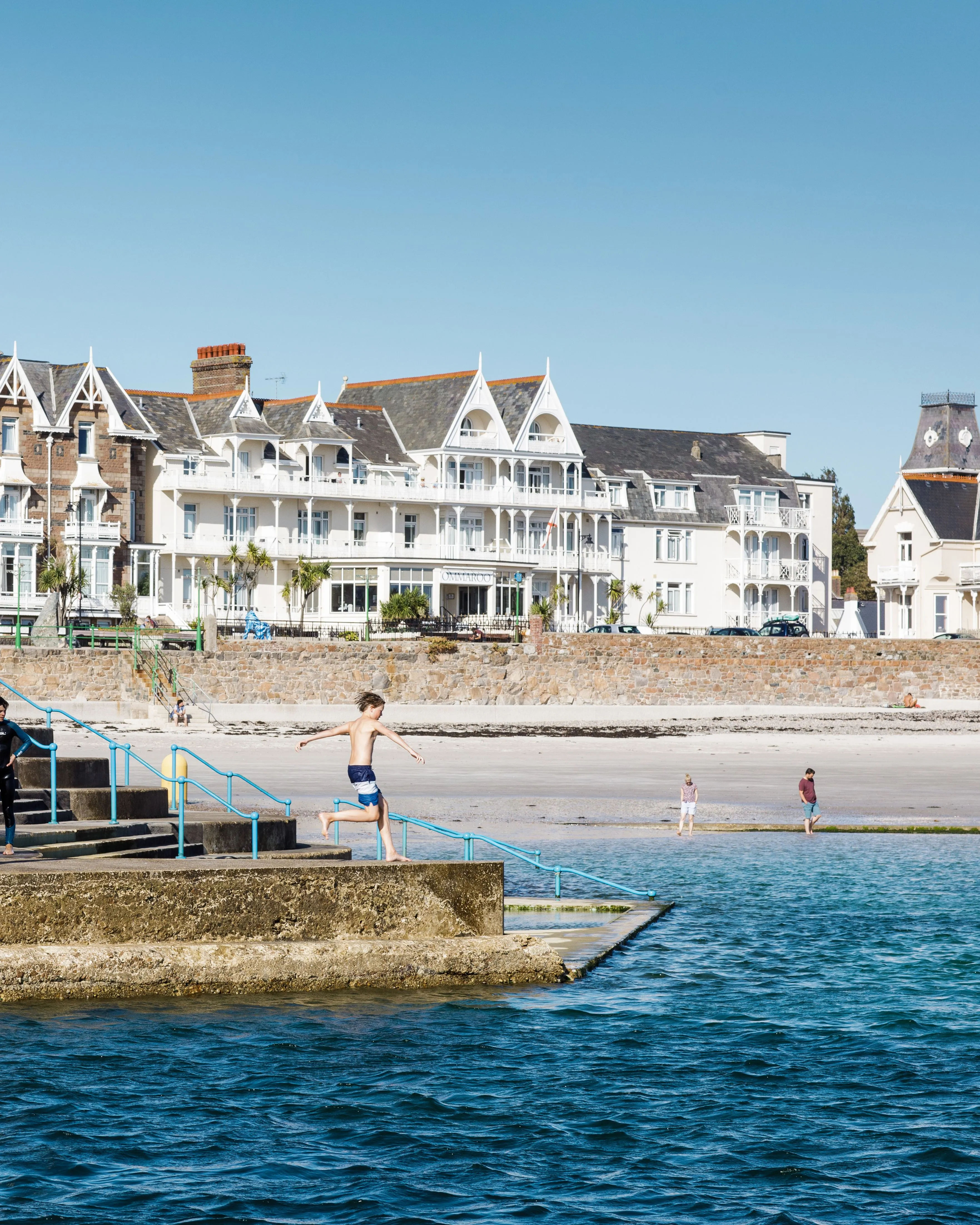 Boy jumping from a seaside harbour wall into clear blue water with beachfront buildings behind