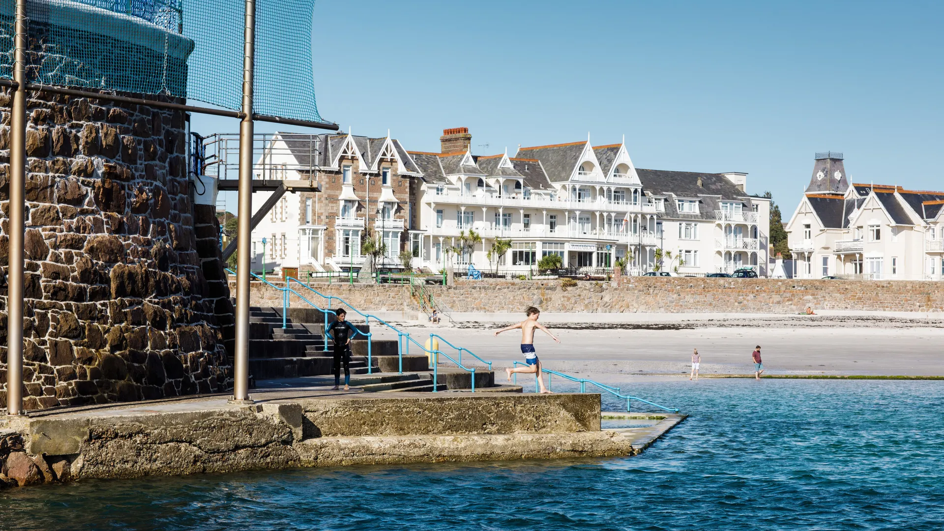 Boy jumping from a seaside harbour wall into clear blue water with beachfront buildings behind