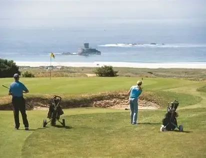 Two golfers on a coastal golf course overlooking the sea, with golf bags on trolleys nearby.