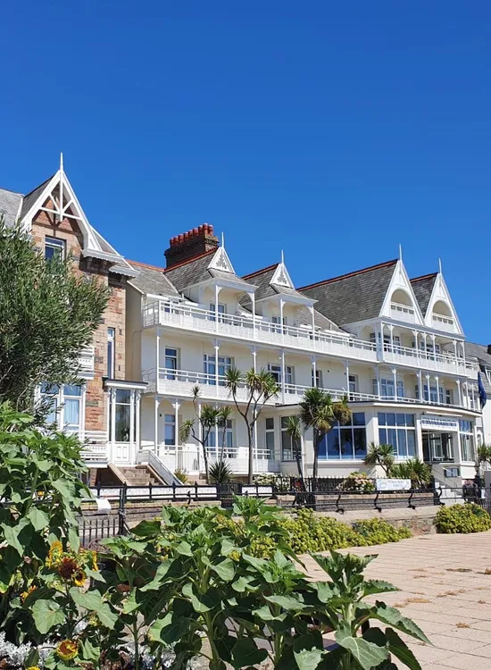 Exterior view of the Ommaroo Hotel with white balconies, gabled rooflines and gardens in the foreground under a clear blue sky.