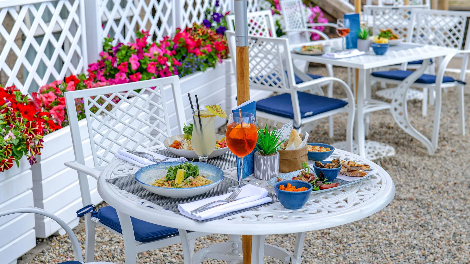 Outdoor food terrace with white tables and chairs, colourful flowers, plated dishes and drinks under parasols.