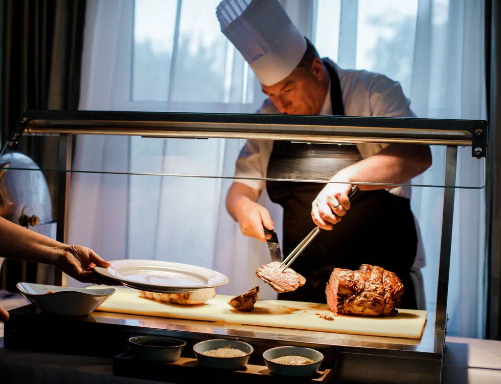 Chef carving roast meat at a serving station while a guest holds out a plate
