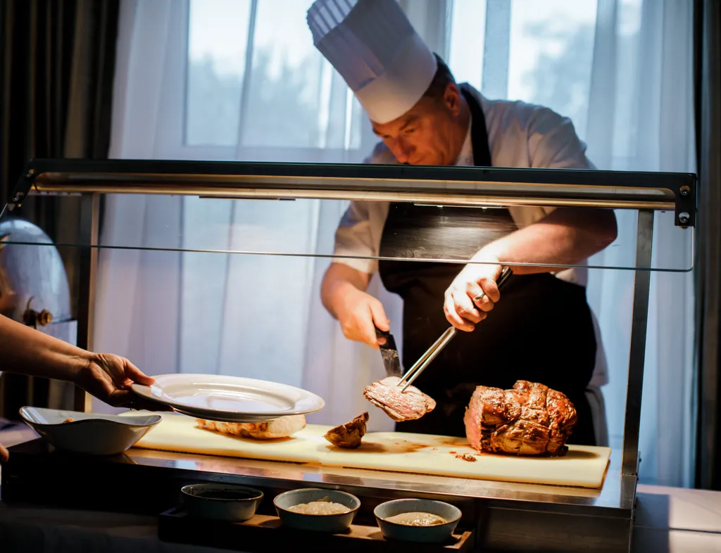 Chef carving roast meat at a serving station while a guest holds out a plate