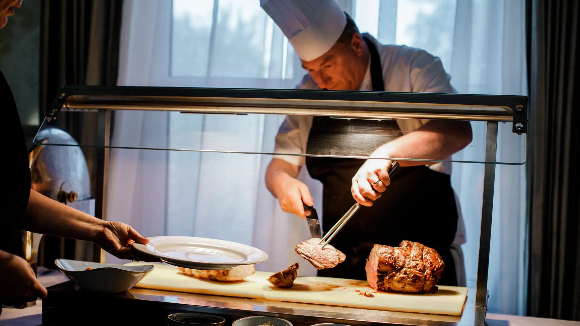 Chef carving roast meat at a serving station while a guest holds out a plate
