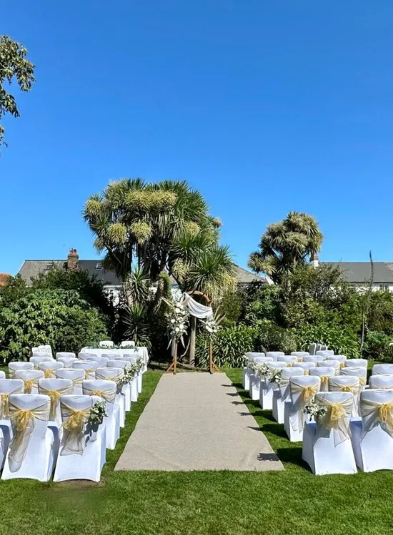 Outdoor wedding ceremony setup in a garden with white chairs, gold sashes and a floral arch