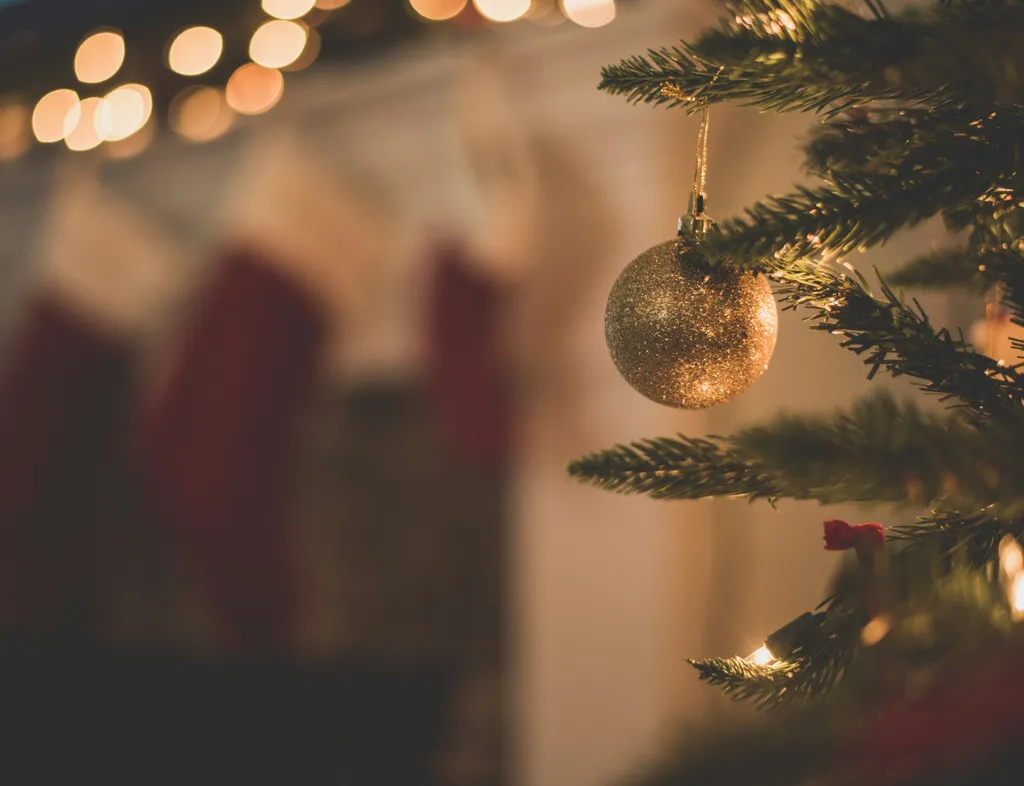 Gold bauble hanging on a Christmas tree with stockings and warm fairy lights in the background