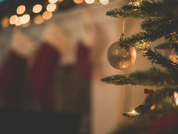 Gold bauble hanging on a Christmas tree with stockings and warm fairy lights in the background
