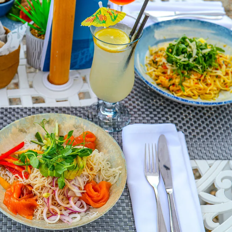 Close-up of a terrace dining table at Ommaroo with fresh noodle dishes, sweet potato wedges, bread, dip and a lemon drink.
