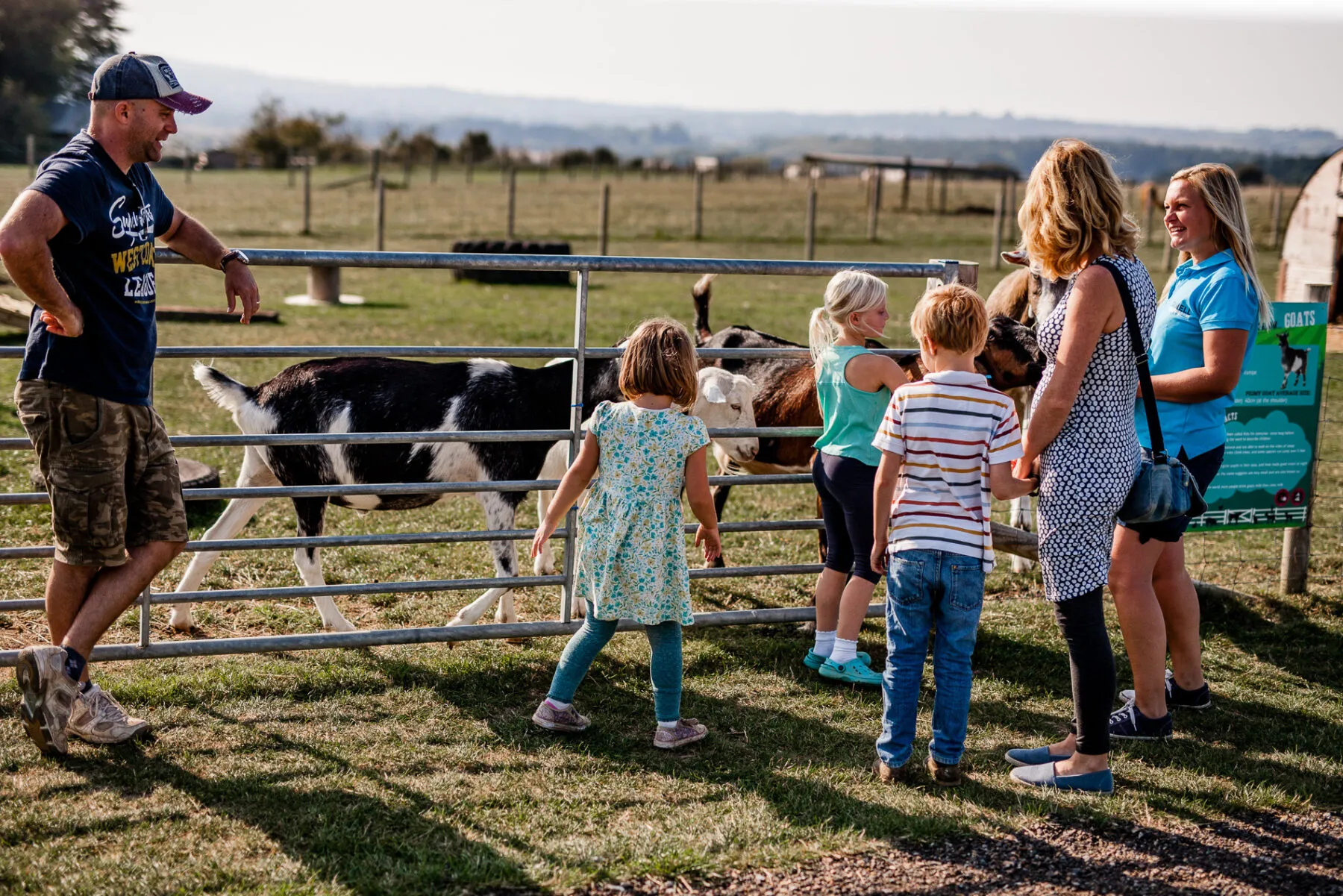 Families and children interacting with goats at a farm park on a sunny day