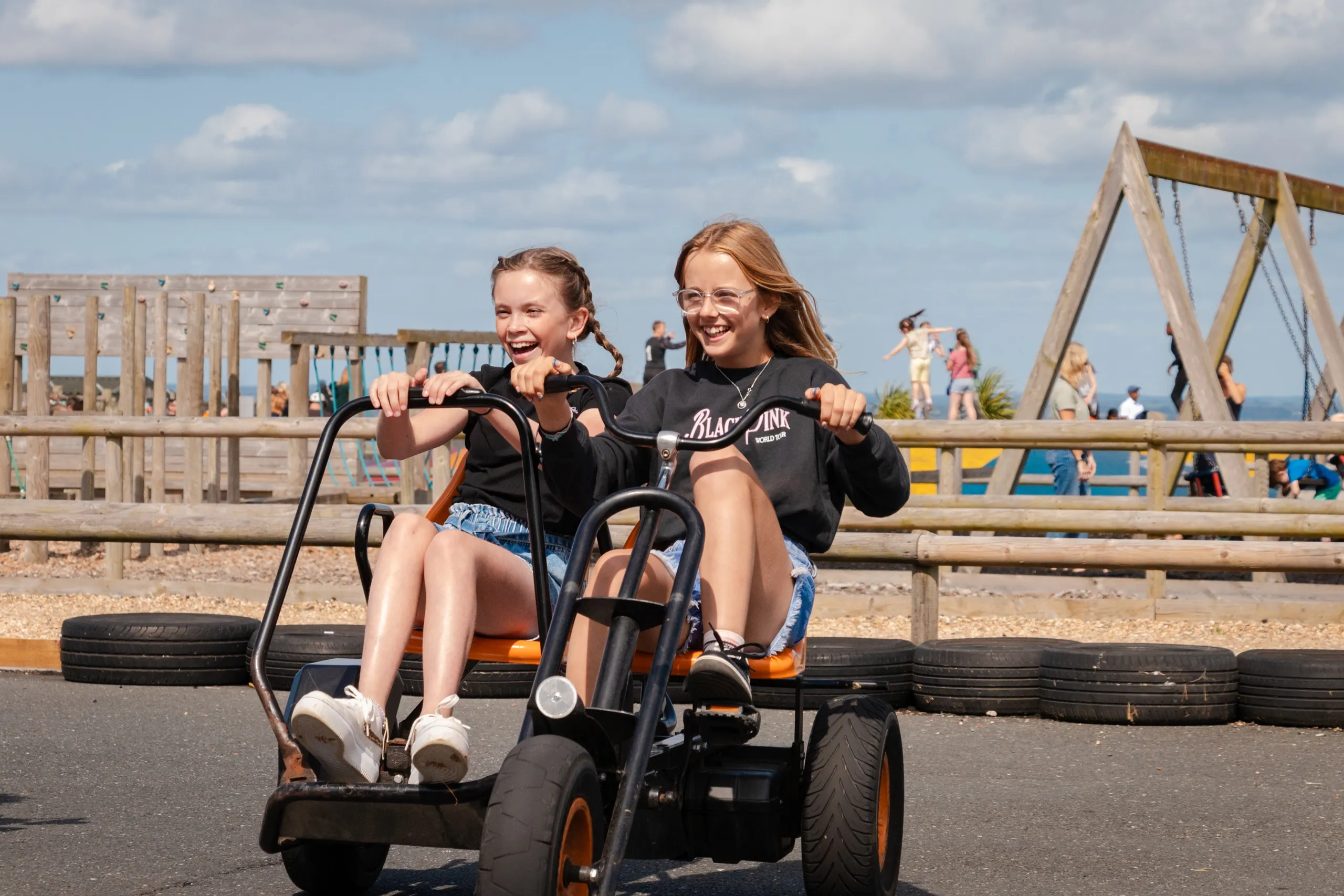Two girls riding a pedal go-kart and smiling at an outdoor park