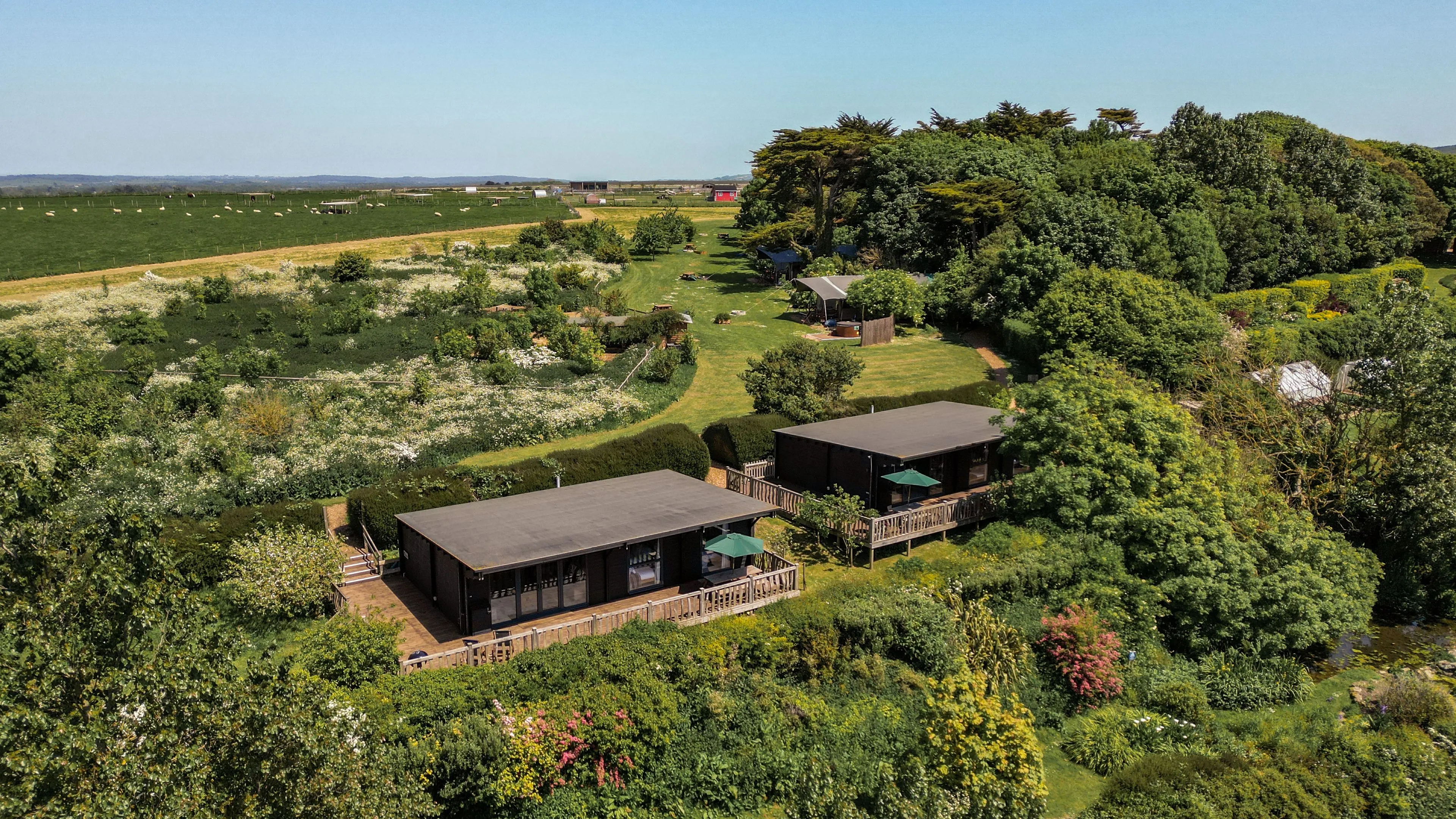 Aerial view of two modern wood cabins with decks amid lush gardens and countryside fields