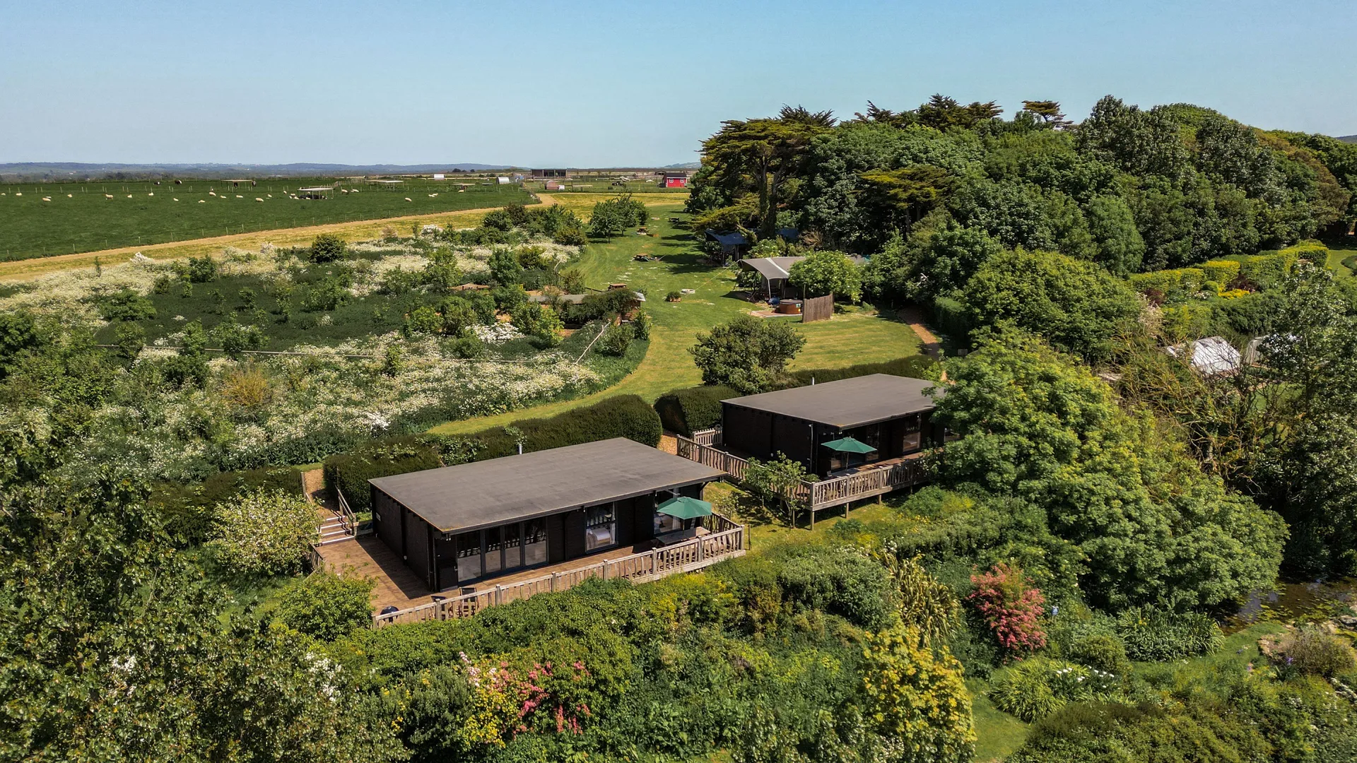 Aerial view of two modern wood cabins with decks amid lush gardens and countryside fields