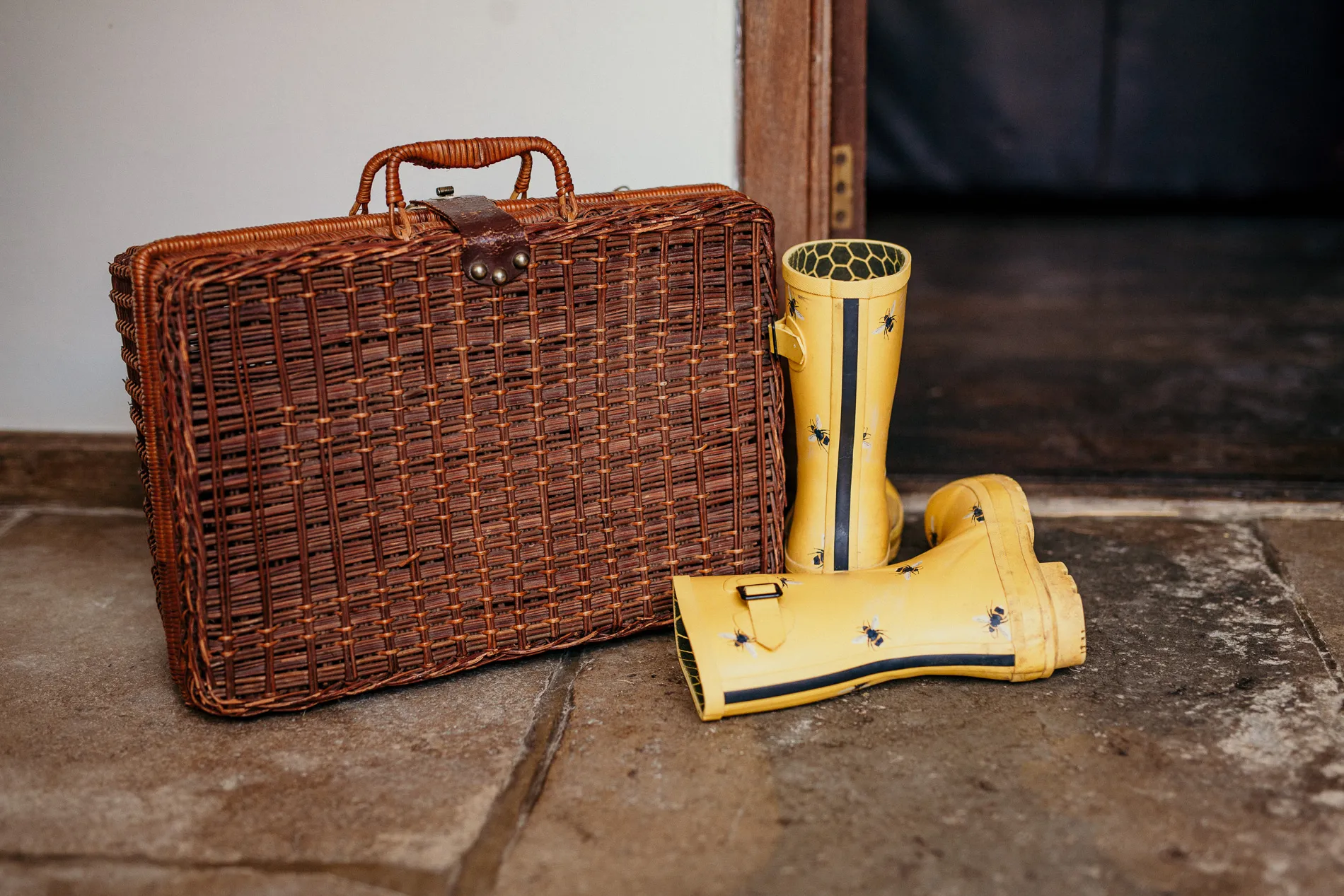 Wicker picnic basket beside yellow wellington boots with bee pattern on a stone floor